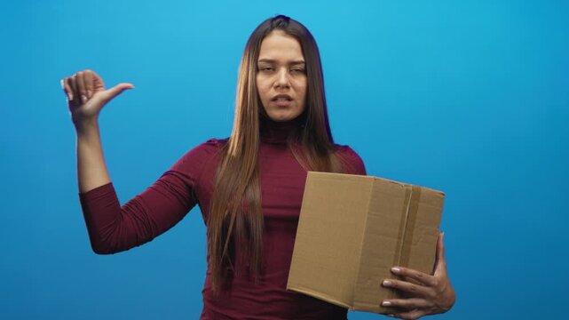 Young brunette woman holding a cardboard box with thumb pointing to self in studio; annoyance delivery complaint.
