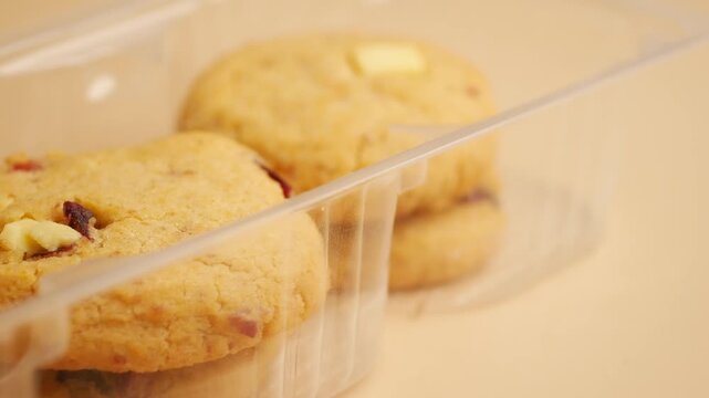 Close up of hand picking up fresh cranberry and white chocolate cookie from plastic container on light background.