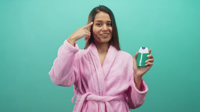 Woman in pink bathrobe points finger to temple while holding toothpaste in studio; playful morning routine.