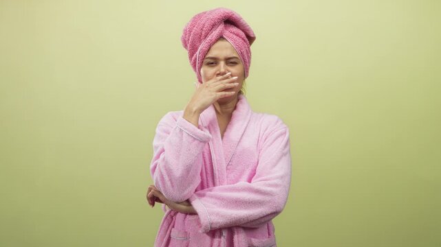 Woman in pink bathrobe and towel turban covering mouth with hand while yawning in studio; sleepiness morning routine.