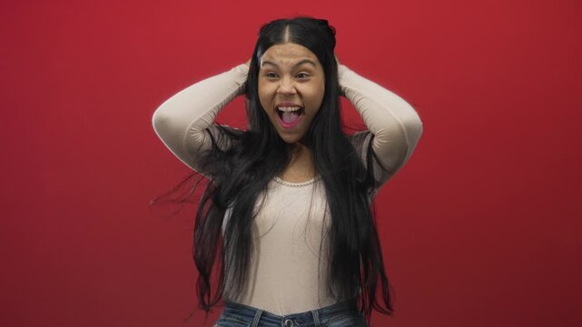 Young latin woman with long hair and open mouth covering ears against red backdrop in studio; surprise.