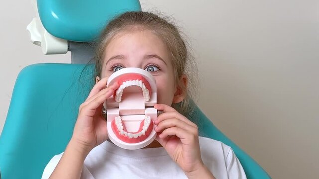 Little girl holding a dental model in front of her mouth while sitting in a dental chair with a light blue background and dental equipment visible. Slow motion footage