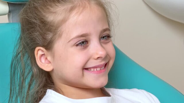 Little girl with light brown hair sits comfortably in a dental chair, smiling in a bright dental office, with dental equipment and a soothing environment visible in the background