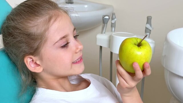 Little girl with long hair sits in a dental chair, taking a bite of a green apple while surrounded by dental equipment and a white sink in a modern clinic. Healthy teeth concept.