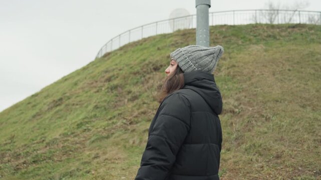 Woman observes lamplight thoughtfully. Female figure pauses to watch lamp thoughtfully outdoors. Caucasian woman in winter attire thoughtfully gazes at lamp amid grassy surroundings