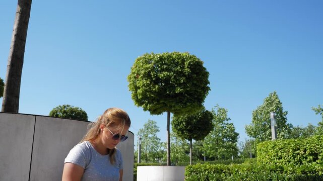 A woman is working on a laptop in a city park. The general layout of a modern park features trimmed bushes, trees, and comfortable benches, on which the woman sits with her laptop.