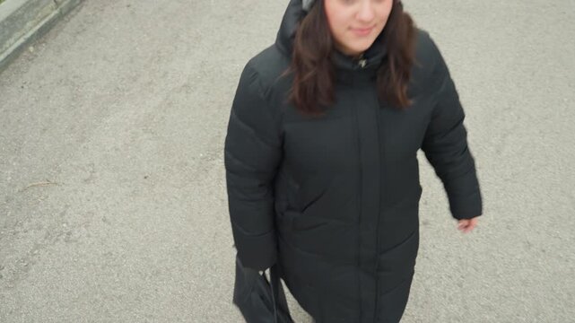 Overhead commuter carrying bag dance rhythm, urban commute footage of woman in grey knit beanie and long black coat walking along sidewalk, steady stride with subtle rhythmic steps, holding black tote
