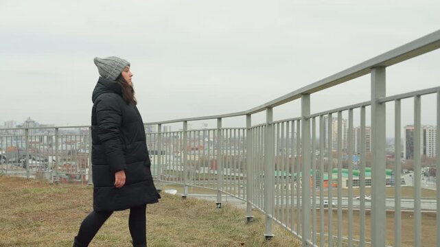Woman walking along rooftop railing city horizon cold breeze and slow purposeful steps in gray beanie and long coat stops to lean and gaze over skyline quiet movement blending with still urban