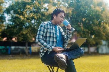 Obraz premium A person is sitting on a folding chair in a park, holding a laptop and a cup, dressed in casual attire with a plaid shirt and blue jeans, enjoying a relaxed outdoor work setting