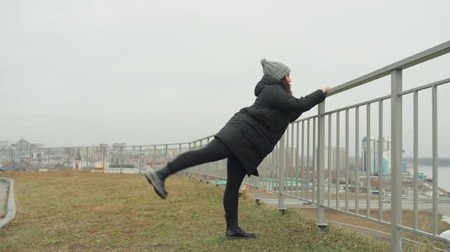 Railside dance improvisation kick skyline, solo performer rehearsing rhythmic leg flicks while holding railing, puffer jacket and beanie against chilly breeze, playful momentum building into balanced