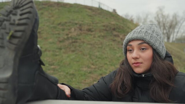 Outdoor dance rehearsal with textured footwear. Knit hat woman inspecting boot sole in park setting. Female dancer preparing playful steps while examining muddy tread on outdoor pathway shoes