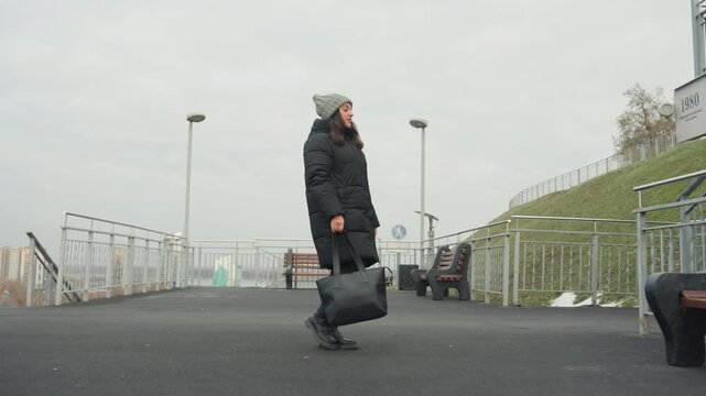 Caucasian woman dancing on waterfront bench, dropping tote bag beside bench, wearing puffer coat and knit hat, energetic improvised spins and steps, expressive smile and animated gestures, interaction