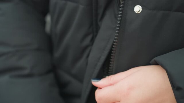 Closeup hands fastening black coat zipper chilly urban street setting subtle swift movements focus on fingers adjusting slider and snaps padded fabric and metal hardware visible everyday preparation