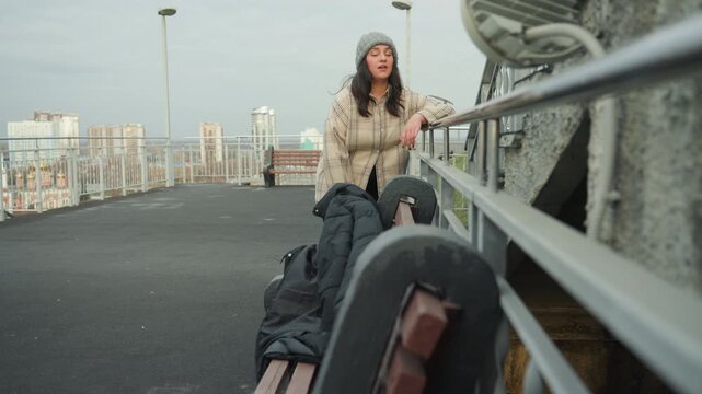 White woman dancing on rooftop walkway with city skyline, beanie and patterned coat, backpack and jacket on bench. Frame arrival as traveler walking across bridge. Frame expressive steps