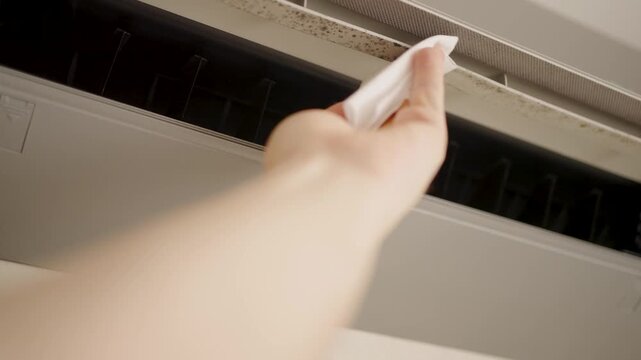 Person Cleaning Moldy Air Conditioner Vent with Wet Wipe. Hand wiping dust and mold from split system air conditioner. Concept of DIY home maintenance, appliance hygiene, improving indoor air quality