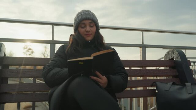 Sunlit woman reading on bench, golden rays creating cinematic flare, focused on worn novel, hands tracing pages, urban hill and railing in background, serene mood, reflective pause, winter light