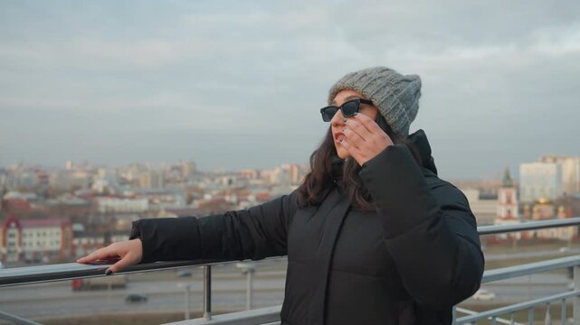 Rooftop winter dance by woman practicing expressive moves overlooking city skyline, knit beanie and sunglasses, padded jacket, calm evening light, solo improvisation with subtle rhythm and relaxed