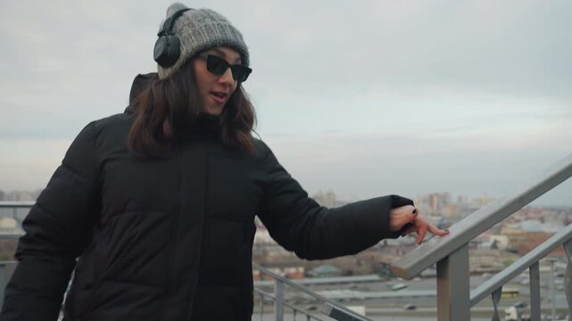 Staircase motion woman dancing with sunglasses and headphones, playful hand gestures while holding railing, pointing toward skyline, cloudy sky bokeh, energetic casual steps, commuter vibe on rooftop