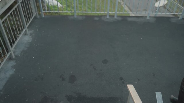 Person in dark jacket walks across small asphalt landing beside metal railing and steps, wearing knit hat under overcast sky brief overhead candid of lone commuter passing camera, wet patches visible