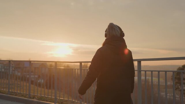 Profile woman with headphones at sunset overlooking railing and city, slow stroll along promenade, warm golden glow, pensive mood and gentle movement, mindfulness vibe and cinematic framing.