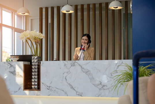 Asian female hotel receptionist answering telephone at luxury front desk in modern boutique hotel lobby with premium hospitality customer service concept  