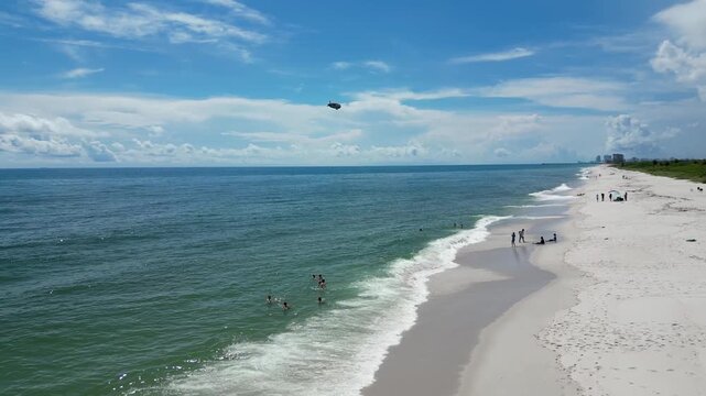 Drone footage of a blimp in a blue sky over the sea shore. People relax on the sand as waves hit the coast. Great for travel and tourism commercials.