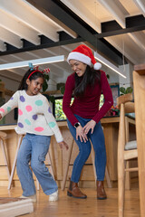 Obraz premium Asian mother and child female dancing in Santa hat and antler headband by wooden kitchen island