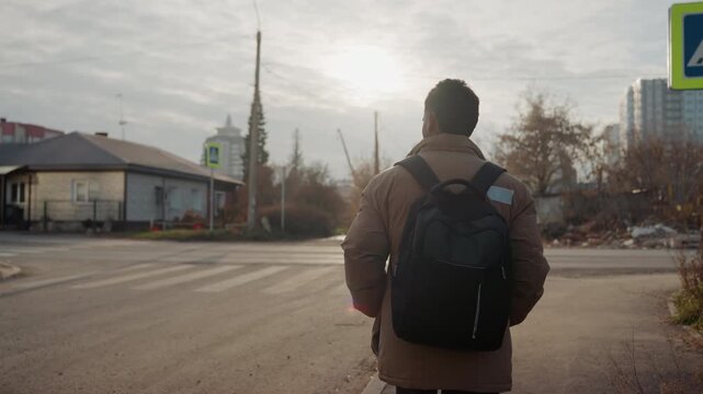 Backpack commuter roadside crossing morning inspects abandoned tire, sun low on horizon, suburban houses and quiet street, back view silhouette, intent to repair flat, hands searching for tools,