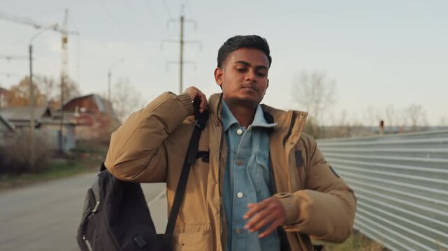Man near metal fence adjusting backpack strap and securing jacket, closeup on hands and shoulders, construction cranes and empty road behind, composed and ready to fix or repair at nearby site