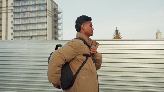 Man adjusting backpack strap by metal construction fence, highrise backdrop and sidewalk, steady determined pace, patched jacket detail visible, candid side profile, city development atmosphere,