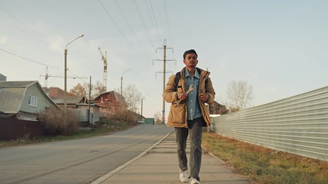 Asian repair technician walking toward camera on suburban road, full stride with backpack and denim layers, corrugated fence and power poles framing scene, late afternoon glow, purposeful approach