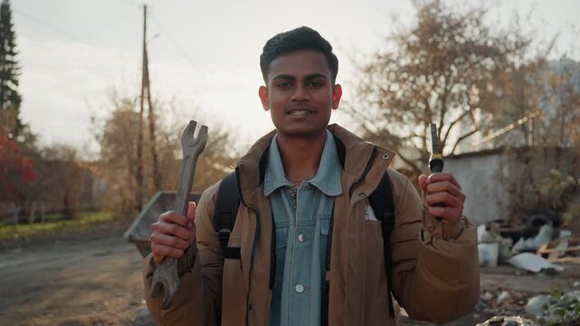 South asian man holding wrench and screwdriver near dumpster, smiling and confident, reclaimed tools and trash pile in background, sunlight backlight, resourceful fixer mood with hopeful tone
