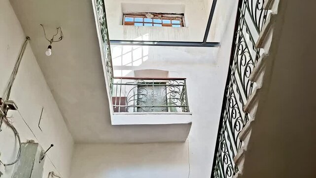 Upward view through multi level atrium with decorative railings, window light, exposed bulb, and shadows, capturing vertical space and architectural perspective