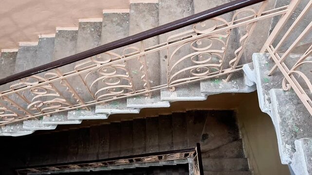 Downward view of square spiral concrete stairwell with decorative metal railings, worn steps, and green discoloration, emphasizing depth, symmetry, and architectural detail
