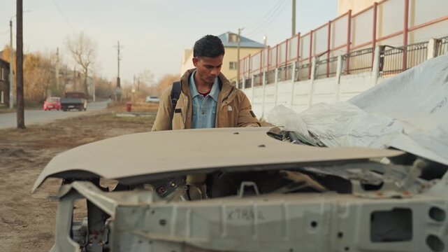 Man examining crushed car hood, checking bolts and panels, photographing damage, calculating estimate, cautious posture, toolkit at side, late afternoon light, fence and houses backdrop, preparing