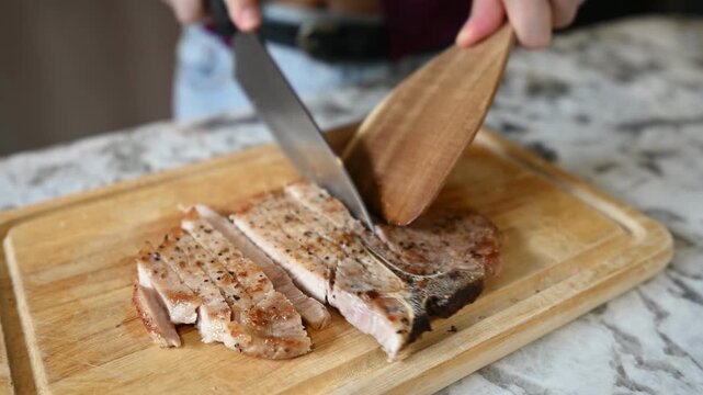 Focused person preparing to cut and slice hot roasted pork chop meat using sharp metal knife upon brown wooden board over granite counter within bright modern home kitchen crafting savory dinner