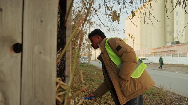 Black painter inspecting roadside wooden fence, performing repair touch up with roller, checking panels and fasteners, urban background with passing pedestrians, focused maintenance action