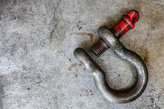 ​A heavy-duty, worn, and rusted industrial screw-pin anchor shackle with a partially faded red-painted pin rests on a scratched metal workshop floor, capturing a detailed texture and rugged feel.