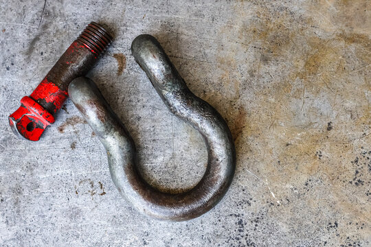 ​A heavy-duty, worn, and rusted industrial shackle and a detached red-painted screw pin rest side-by-side on a scratched metal workshop floor, capturing detailed texture and a rugged feel.