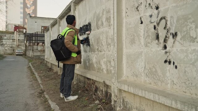Black municipal worker cleaning graffiti wall, reflective vest and backpack, using scraper and solvent to remove spray paint from concrete fence in quiet urban alley during daytime, focused on repair