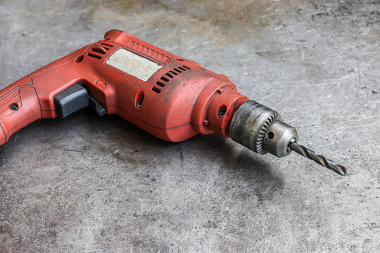 Used red corded power drill with metal chuck and drill bit on a grimy steel workshop table surface. Isolated shot of durable, messy hand tool showing wear and tear for heavy construction work.