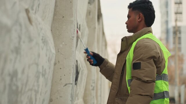 Closeup black worker repairing concrete wall with small roller and brush, wearing highvisibility vest and heavy coat, gloved hands applying patching compound to damaged surface, urban backdrop