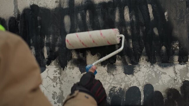 Closeup roller covering black graffiti, south asian worker repairing concrete wall, wearing high visibility vest and gloves, methodical horizontal strokes over cracked textured surface, primer