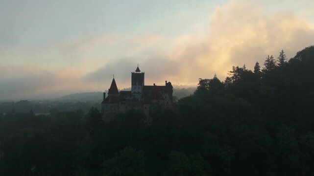 Stunning ascending aerial view rising high above the iconic Bran Castle through moody thermal inversion mist and dense low clouds at majestic summer sunrise, cinematic establishing drone shot, graded