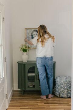 Woman hanging framed artwork above green console