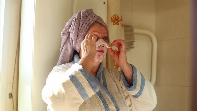 A woman in a robe and towel wrapped hair carefully removes a sheet mask in front of a bathroom mirror, soft light, gentle motion, and quiet self care define the moment.