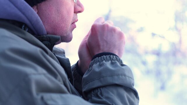 A man blows warm breath onto his cupped hands in a snowy forest, close-up of resilience, quiet ritual, and winters chill met with simple, human warmth.