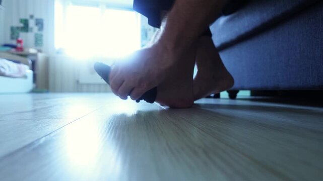 Low angle shot of a man putting on black socks while seated, bare feet on light wood floor, sofa and table blurred behind, capturing quiet daily ritual, comfort, and mindful preparation.