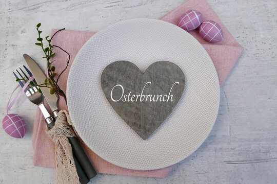 Osterbrunch Gedeck f&uuml;r Gastronomie und Catering-Marketing &ndash; Gedeckter Tisch mit Schriftzug Osterbrunch auf einem Herz auf rosa Stoffserviette, daneben Besteck mit Spitzenband und dekorative Ostereier,