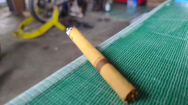 Close up of a lit cigarette burning with smoke wafting on a green table. Traditional Indonesian clove cigarette (kretek) burning and producing smoke.